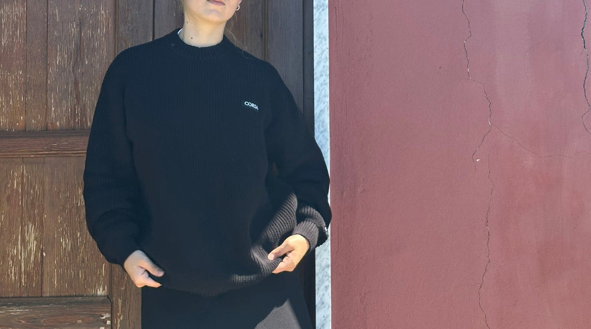Person wearing a black sweater with a logo, standing against a textured wall.