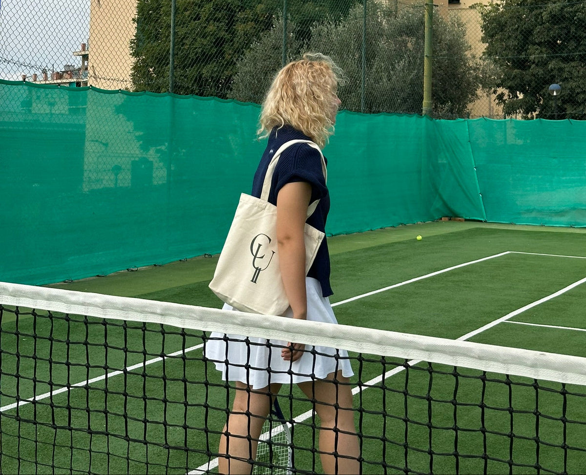 Person standing on a tennis court with a green fence and building in the background